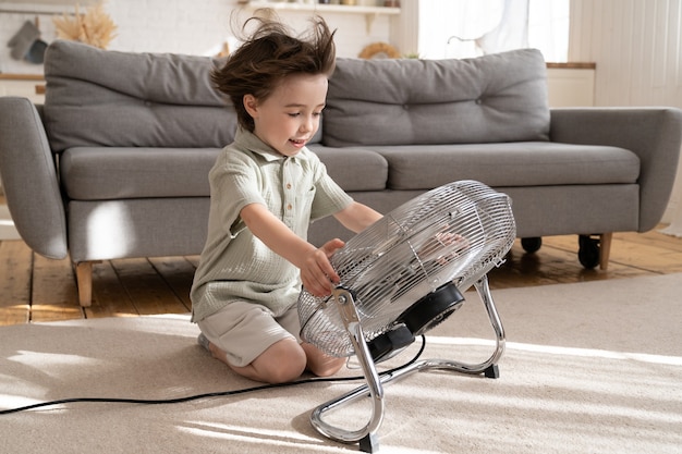 Menino alegre refrescando com grande ventilador em casa sozinho menino pequeno desfruta do forte vento fresco