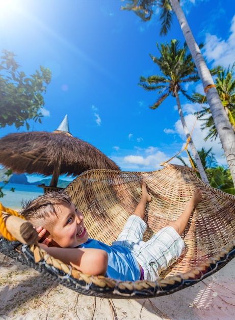 Menino alegre relaxando em uma praia tropical na rede.