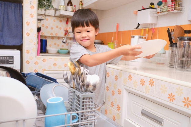 Foto menino asiático bonito se divertindo lavando a louça carregando a máquina de lavar louça na cozinha em casa pequenas tarefas de ajudante doméstico para crianças conceito de desenvolvimento de habilidades de funcionamento executivo