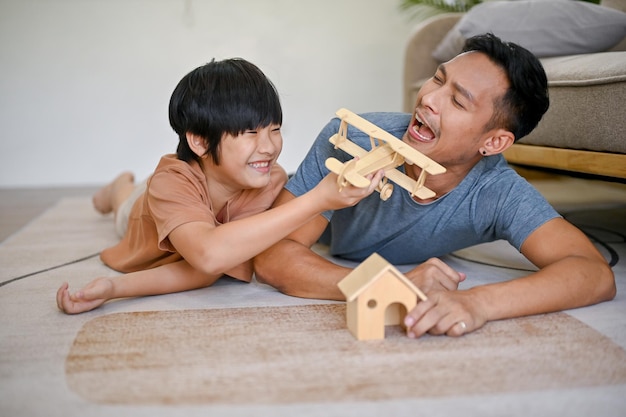 Menino asiático brincalhão jogando um modelo de figura de madeira com seu pai enquanto estava deitado no chão