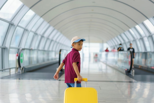 Menino asiático com mala amarela, camisa e calções a caminhar no terminal do aeroporto.