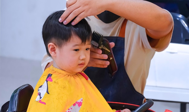 Menino asiático de 2 anos de idade em cadeira alta em forma de carro de brinquedo com corte de cabelo na barbearia infantil