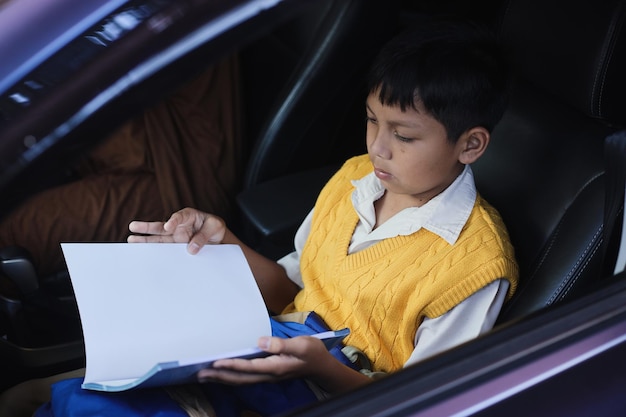 Menino asiático de uniforme está sentado em um carro e lendo um livro quando vai para a escola