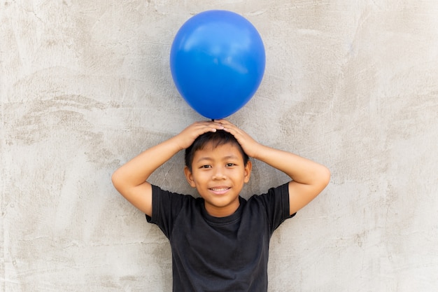 Menino asiático pequeno que guarda o balão sobre sua cabeça com fundo cinzento.