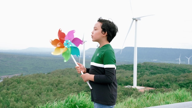 Menino asiático progressivo brincando com brinquedo de turbina eólica no campo verde da fazenda de turbinas eólicas sobre a colina Energia verde de gerador eólico elétrico renovável