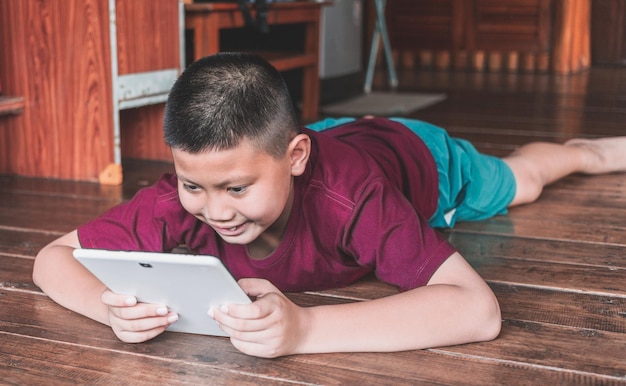 Menino asiático sorridente sentado no chão de madeira e jogando em um tablet digital em casa