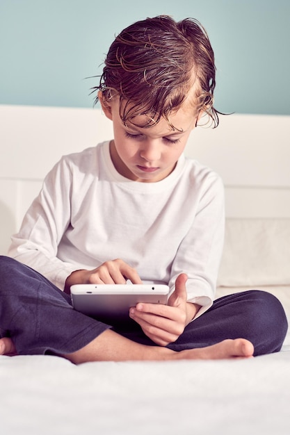 Menino atento com o cabelo molhado sentado na cama e jogando jogo no tablet no quarto