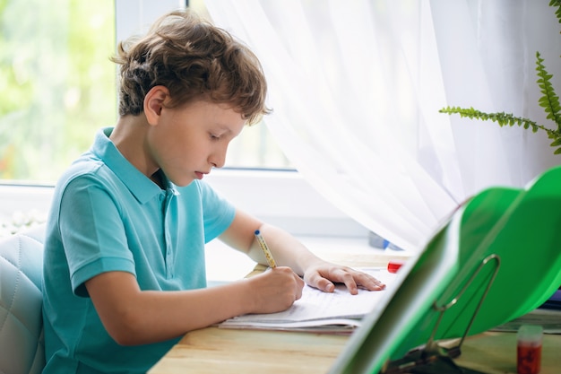 Menino atento, escrevendo no caderno enquanto está sentado na mesa e fazendo lição de casa.