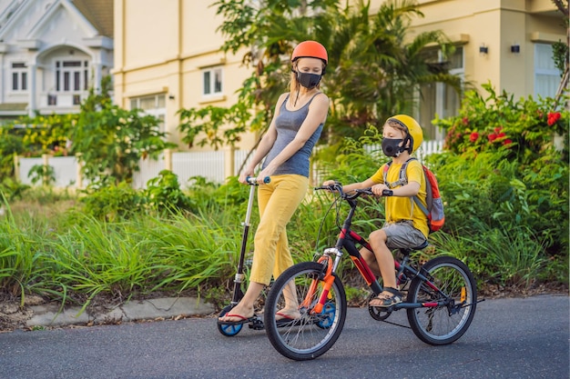 Menino ativo da escola e sua mãe com máscara médica e capacete de segurança andando de bicicleta com mochila