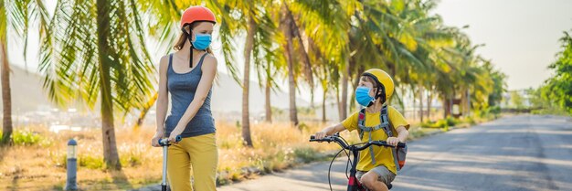 Foto menino ativo da escola e sua mãe com máscara médica e capacete de segurança andando de bicicleta com mochila