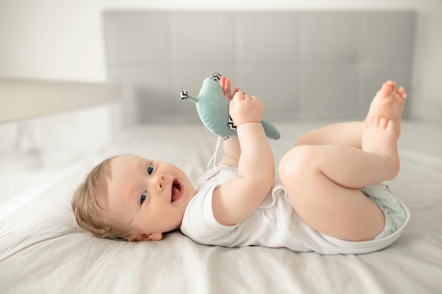 Foto menino bebê fofo usando fralda brincando com os pés na cama em um quarto ensolarado