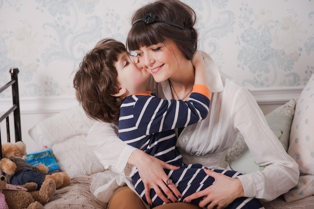 Menino beija sua mãe e abraços. mulher jovem feliz se divertir com o menino bonito criança. mãe e filho sentam-se na cama antes de irem para a cama. conceito de boa noite