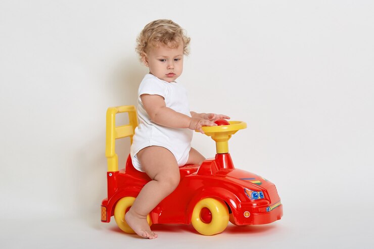 Foto menino bonitinho andando em um carrinho de brinquedo vermelho e amarelo, olhando para longe, posando descalço, veste macacão