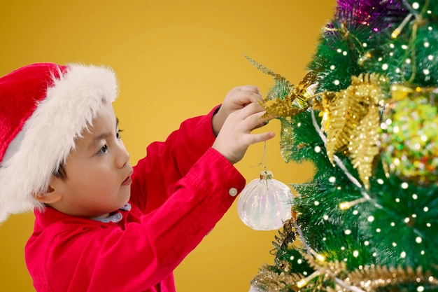 Menino bonitinho com chapéu de Papai Noel pendurando uma bola de Natal na árvore de Natal em casa