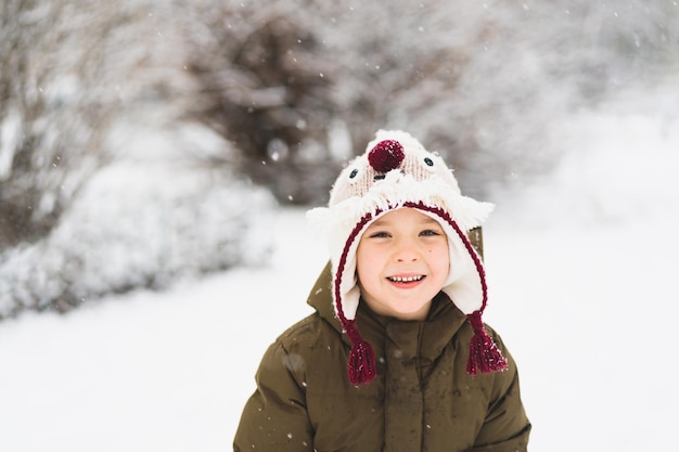 Menino bonitinho com chapéu engraçado de inverno caminha durante uma queda de neve ao ar livre atividades de inverno para crianças