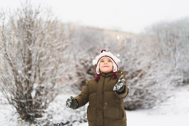 Menino bonitinho com chapéu engraçado de inverno caminha durante uma queda de neve ao ar livre atividades de inverno para crianças