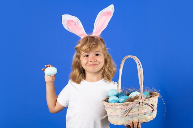 Menino bonitinho com orelhas de coelho segurando ovo de páscoa na cor de fundo azul Feliz feriado da páscoa Criança bonitinha usando orelhas de coelho isoladas em fundo azul