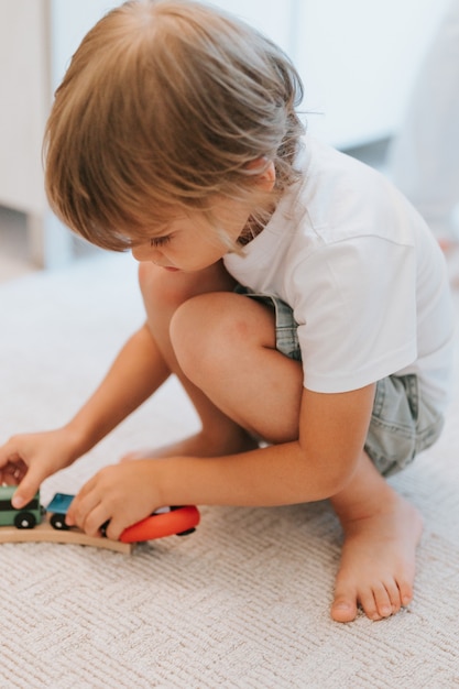 Menino bonitinho de cinco anos de idade em uma camiseta branca, brincando com uma ferrovia de madeira e trens de brinquedo no chão, no tapete da sala. crianças brincam em casa