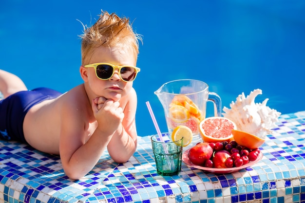 menino bonitinho de óculos escuros e maiô bebe limonada à beira da piscina.