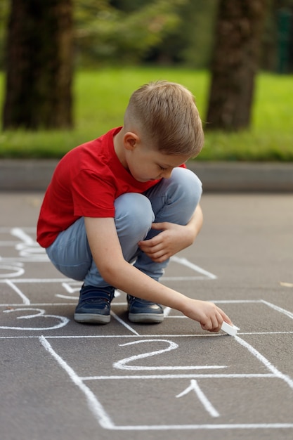 Menino bonitinho desenhando amarelinha com giz no playground. Divertido jogo de atividades para crianças no playground do lado de fora.