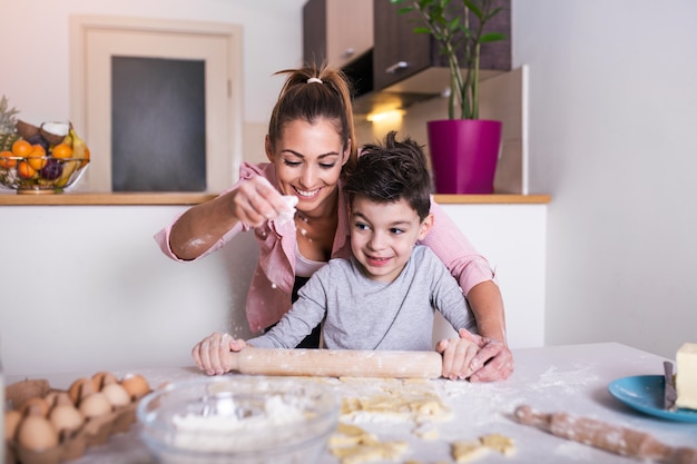 Menino bonitinho e sua linda mãe de avental estão sorrindo enquanto amassar a massa na cozinha