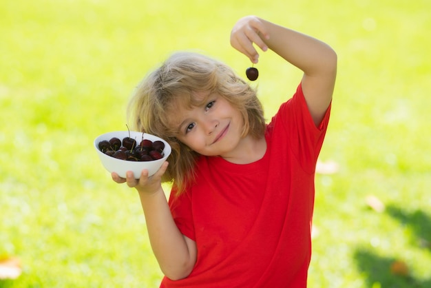 Menino bonitinho em camiseta vermelha comendo cerejas fazendo caretas e brincando com as cerejas se divertindo