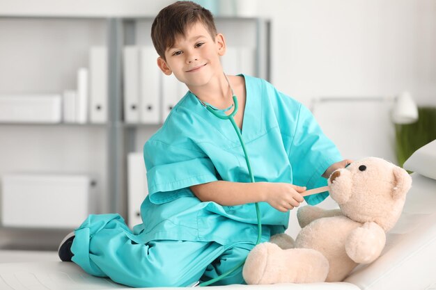 Menino bonitinho em uniforme de médico brincando com urso de brinquedo no hospital