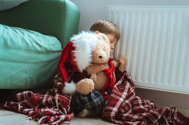 Menino bonitinho embrulhado id plaid sentado pelo aquecedor com ursinho de pelúcia vestido com chapéu de natal