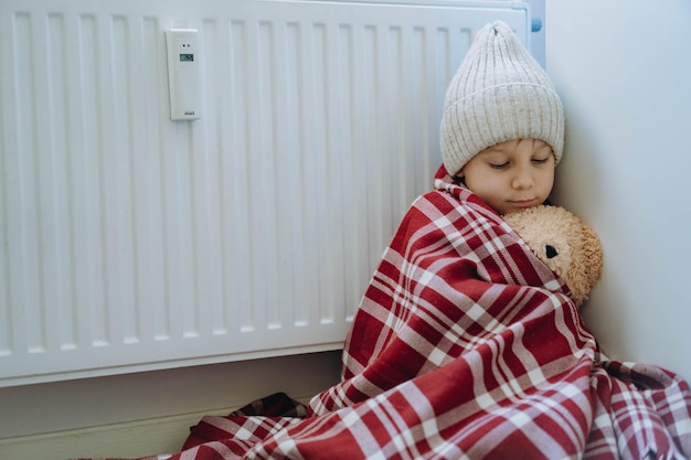 Menino bonitinho embrulhado id plaid usando chapéu de malha sentado perto do aquecedor abraçando o ursinho frio em casa