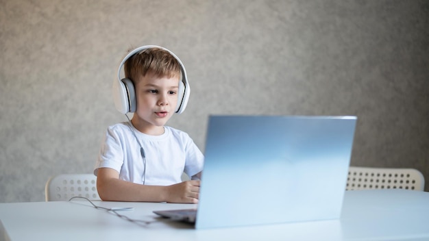 Menino bonitinho está assistindo desenhos animados ou um filme em um laptop Criança caucasiana senta-se à mesa em casa e ouve música em fones de ouvido