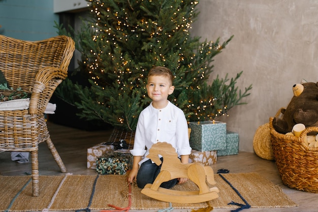 Menino bonitinho está sentado perto de uma árvore de Natal e um cavalo de balanço de madeira vestindo uma camisa branca