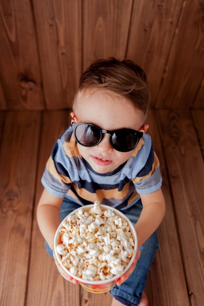 Menino bonitinho, menino, 23 anos, óculos de cinema 3d, segurando balde para pipoca, comendo fast-food em fundo de madeira Conceito de estilo de vida infantil para crianças Copiar espaço