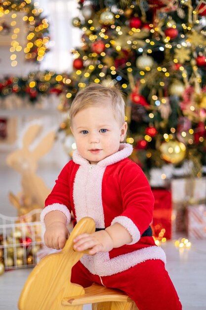 Menino bonitinho na fantasia de Papai Noel monta um brinquedo de balanço sob uma árvore de Natal festiva com presentes na sala de estar da casa um grande retrato de ano novo do bebê