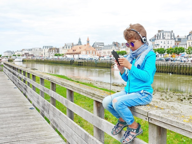 Menino bonitinho ouve música com fones de ouvido. ele adora música. ele se senta em uma ponte com um rio e telhados ao seu redor