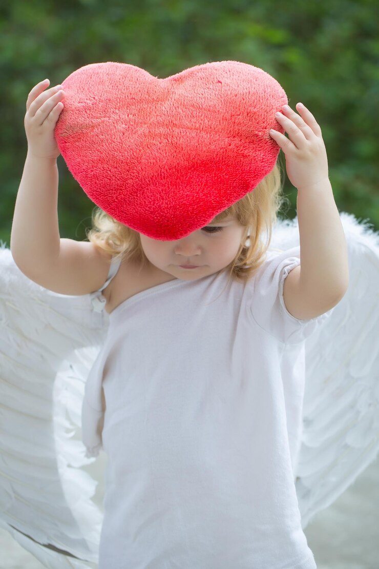 Foto menino bonitinho pequeno com cabelo comprido loiro em asas de anjo de penas brancas e pano segurando travesseiro de coração vermelho ao ar livre em fundo verde natural