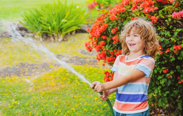 Menino bonitinho regando flores no jardim no dia de verão criança usando mangueira de jardim água engraçada