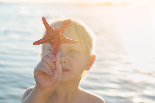 Menino bonitinho segurando estrela do mar em sua mão minúscula contra o mar e o sol