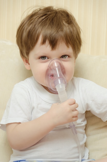 Menino bonitinho sentado em casa no sofá e respirar através de um inalador de nebulizador.