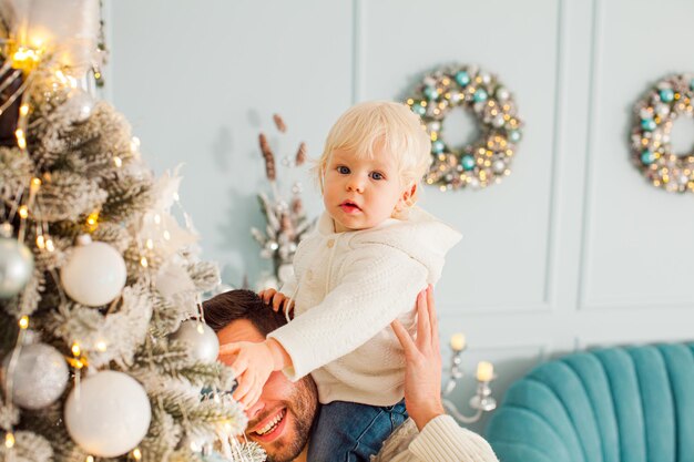 Menino bonitinho sentado nos ombros de seus pais pendurando brinquedos de Natal em uma árvore olhando para a câmera Sala com decoração moderna e elegante em um fundo