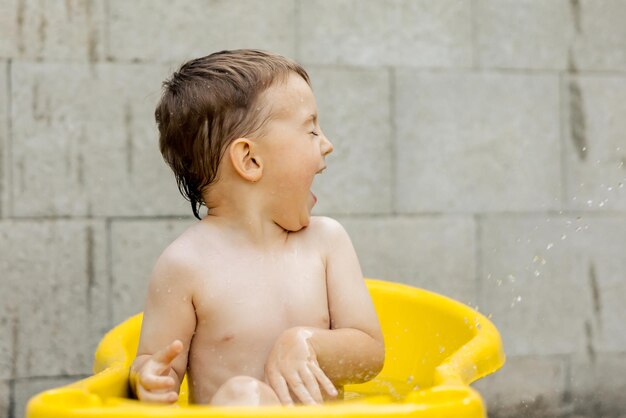 Menino bonitinho tomando banho na banheira amarela ao ar livre Criança feliz está espirrando brincando com água e se divertindo Temporada de verão e recreação Ficando fresco no calor do verão Diversão de água no quintal