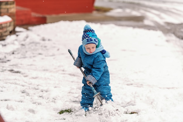 Menino bonitinho vestido com roupas de inverno azul brincando com garfo de feno na neve.