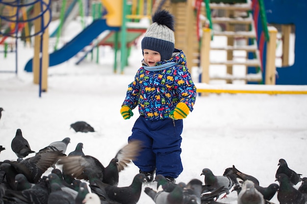 Menino bonito alimenta pássaros no parque de neve de inverno ao ar livre