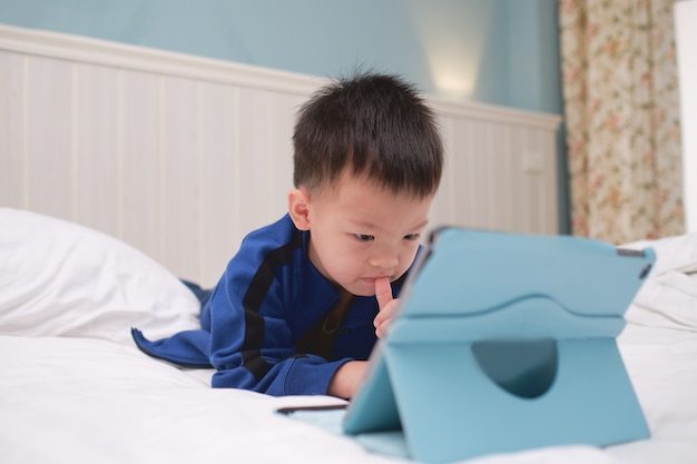 Menino bonito asiático de 3 a 4 anos de idade sorrindo enquanto joga, assistindo desenhos animados, usando o computador tablet pc, crianças viciadas em gadgets, learning tablet for kids, conceito de brinquedo educacional para crianças