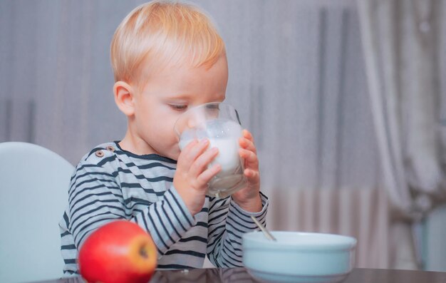 Menino bonito bebê tomando café da manhã. Nutrição do bebê. Comer saudável. Criança fazendo lanche. Nutrição saudável. Beber leite. Criança segura copo de leite. Garoto bonito garoto sente-se à mesa com prato e comida. Comida saudável