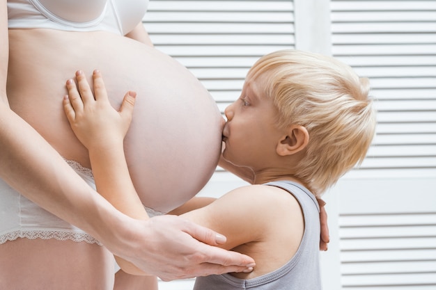 Menino bonito, beijando e abraçando a barriga da mãe grávida. Segundo conceito de gravidez.