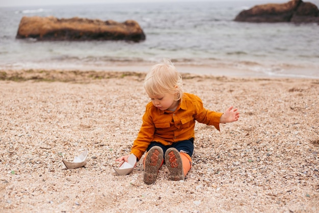 Menino bonito brincando com barquinho de papel na areia no outono