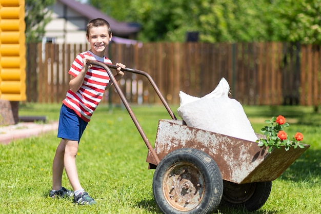 Menino bonito brincando com um grande carrinho de mão velho no quintal no jardim ao ar livre