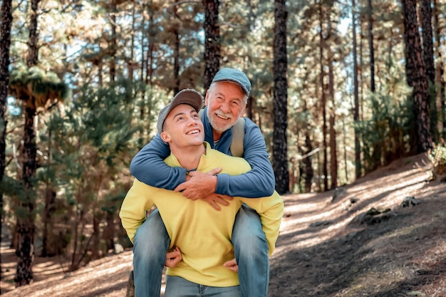 Menino bonito carrega seu avô nos ombros enquanto ele caminha feliz na floresta Casal sorridente de várias gerações desfrutando da montanha e da natureza juntos A nova geração ajuda a antiga