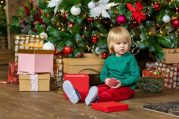 Menino bonito com fantasia de Natal debaixo da árvore com o presente. criança brincando com bola, brinquedo em casa. criança está esperando ano novo, papai noel