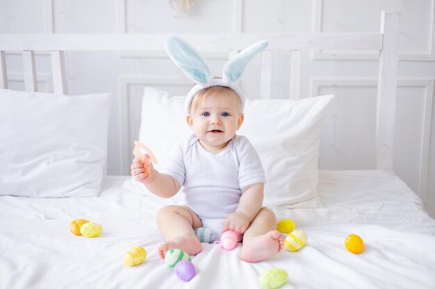 Menino bonito com orelhas de coelho e ovos coloridos em uma cama branca em casa brincando ou comendo ovos bebê loiro conceito de feliz páscoa